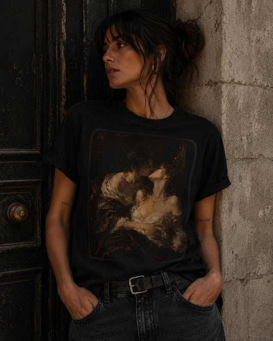 A woman wearing a black graphic t-shirt with a classical painting print, leaning against a stone wall next to a dark wooden door.