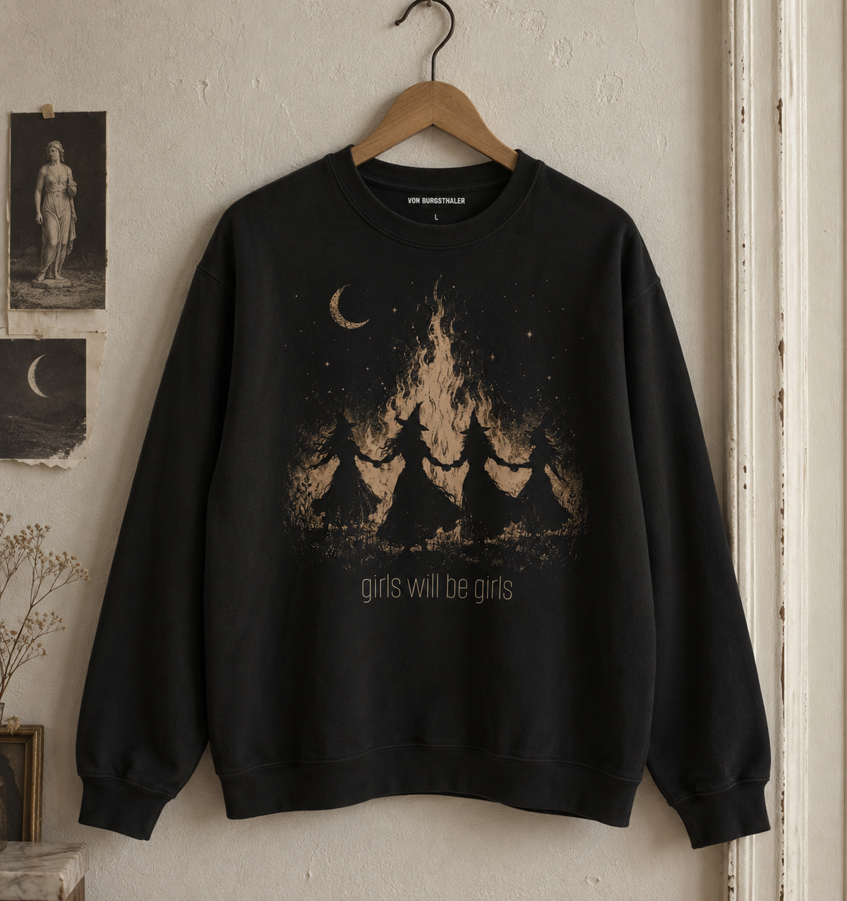A black crewneck sweatshirt hanging on a wooden hanger against a textured beige wall. The sweatshirt features a graphic print of four girls holding hands in front of a campfire under a crescent moon, with the text 'girls will be girls' printed below.